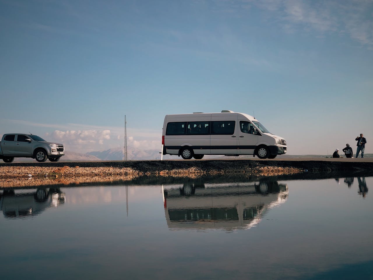 mobile-02 A van and trucks parked by a tranquil lakeshore in Adıyaman, Turkey, beautifully reflected in still water.